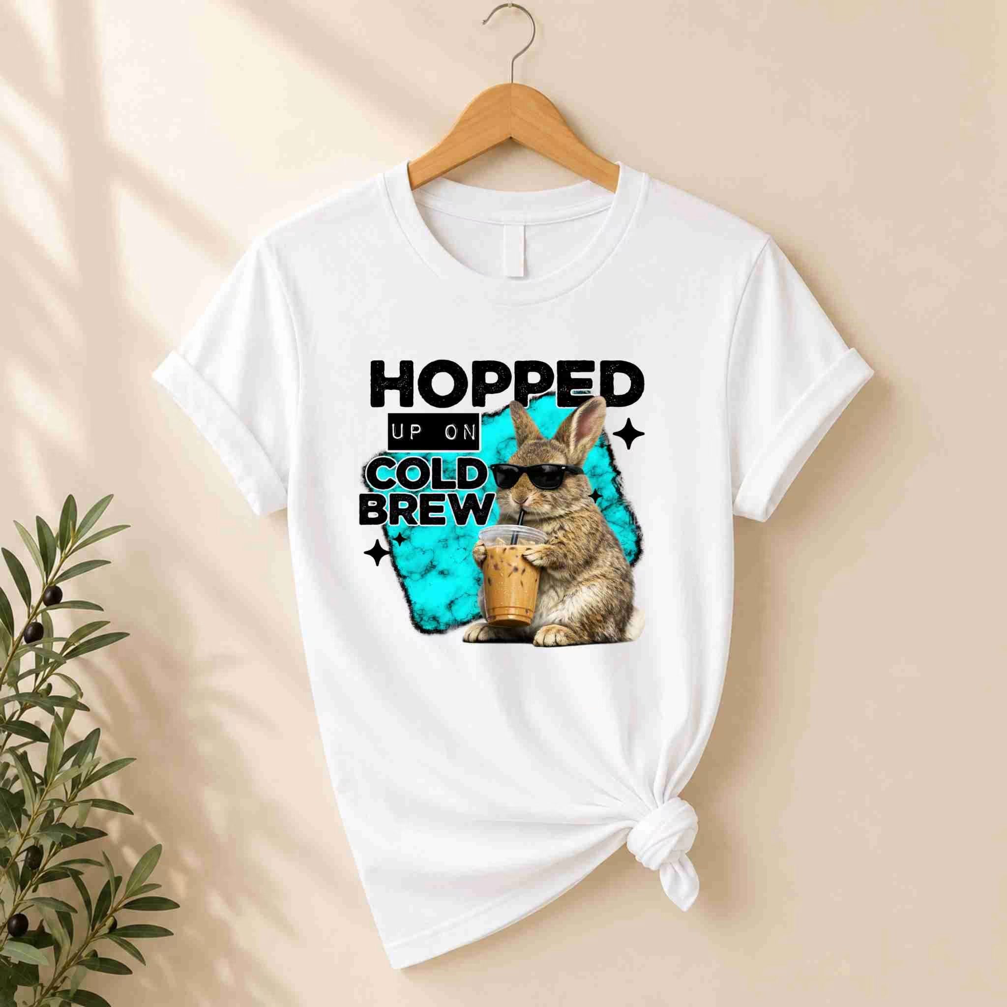 The "Hopped Up On Cold Brew Bunny Shirt" by Becca's Banging Designs LLC is a white graphic tee with a cool bunny holding iced coffee, sunglasses on, and the phrase “Hopped Up On Cold Brew” above. A leafy plant rests beside the shirt on a wooden hanger.