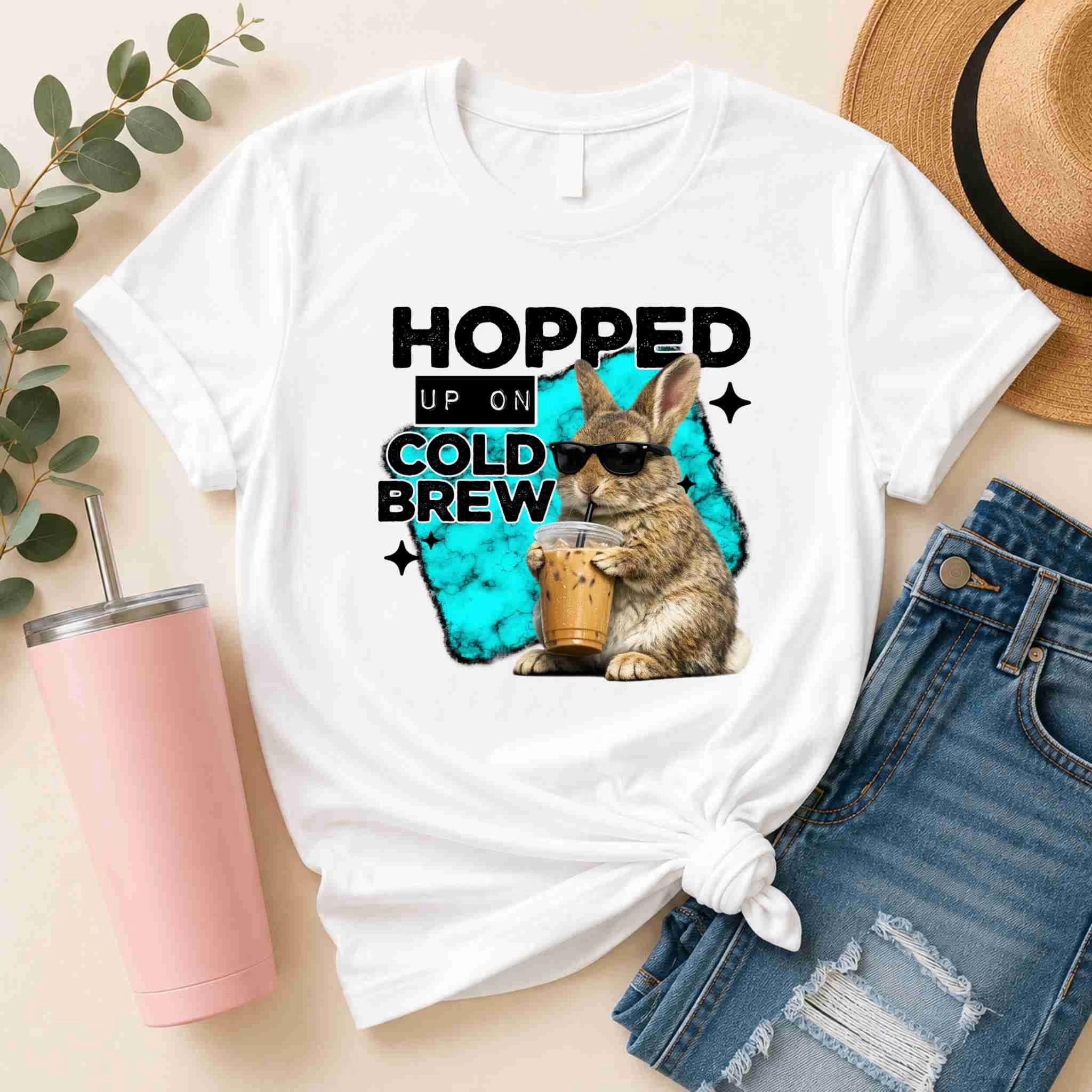 The Hopped Up On Cold Brew Bunny Shirt by Becca's Banging Designs LLC is a funny coffee graphic tee with a sunglasses-wearing bunny and "Hopped up on cold brew" text—perfect for casual style with denim shorts and a straw hat.
