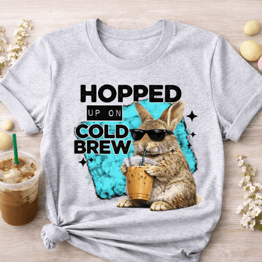 Gray t-shirt with a rabbit drinking iced coffee and 'Hopped Up on Cold Brew' text on a wooden surface.