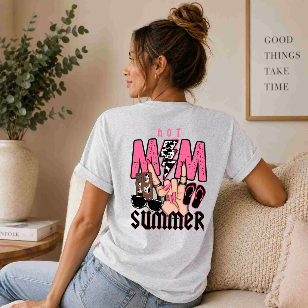 A woman with a ponytail sits on a sofa, viewed from behind, wearing the Becca's Banging Designs LLC Hot Mom Summer Tee – Trendy Mama Graphic Shirt. A plant and framed sign create a cozy vibe. Perfect for fans of women's graphic tees.