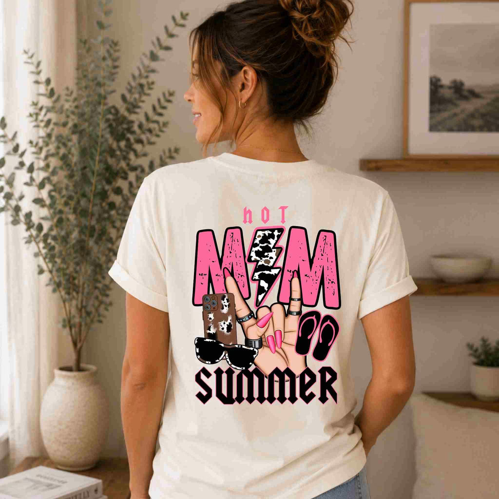 A woman with a messy bun wears the Hot Mom Summer Tee – Trendy Mama Graphic Shirt by Becca's Banging Designs LLC, featuring sunglasses, nails, and sandals graphics on the back. She stands indoors facing away near a shelf and potted plant.