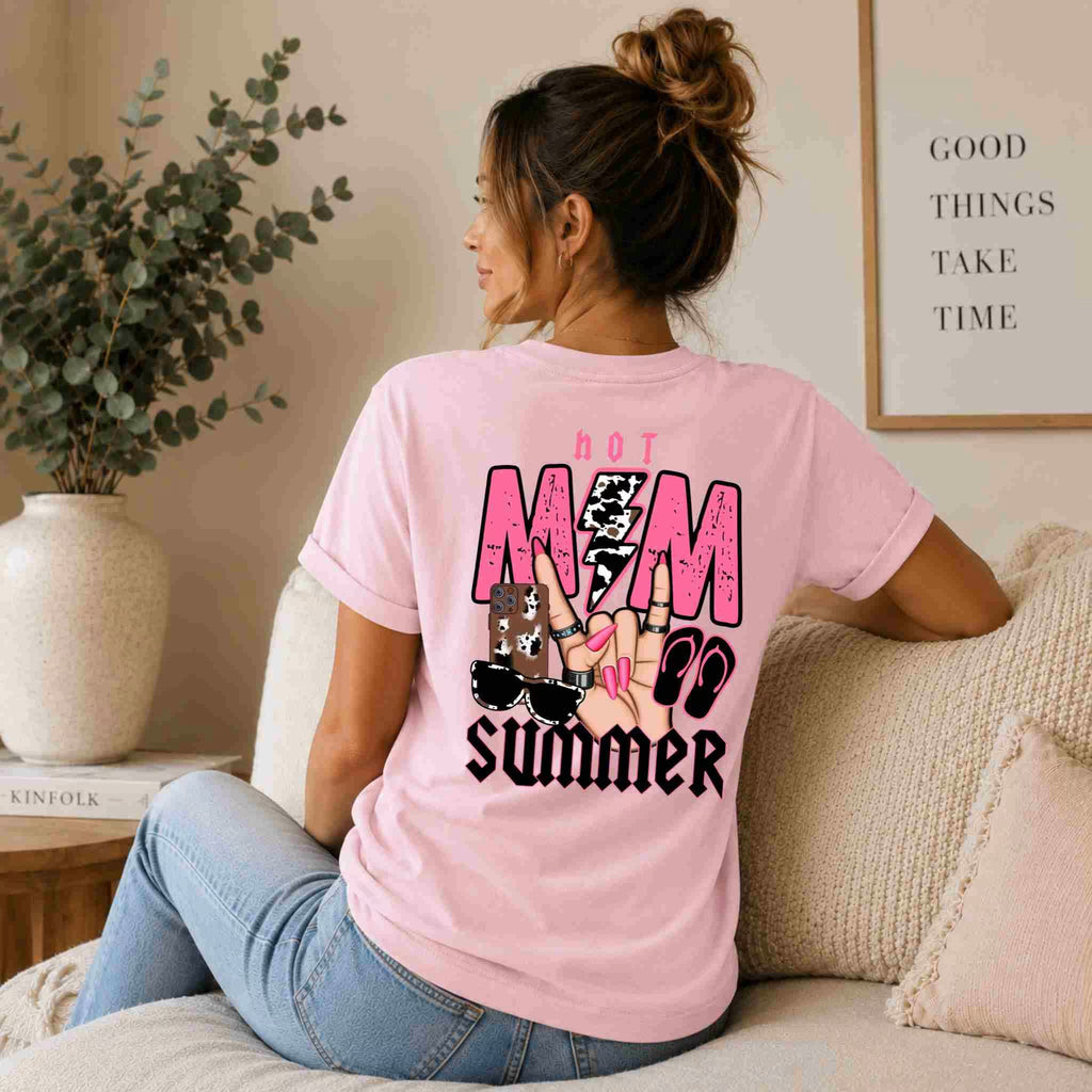 A woman with a ponytail relaxes on a couch, wearing Becca's Banging Designs LLC's Hot Mom Summer Tee – Trendy Mama Graphic Shirt, as she glances beside a framed “Good Things Take Time” sign—ideal for fans of bold women's graphic tees.