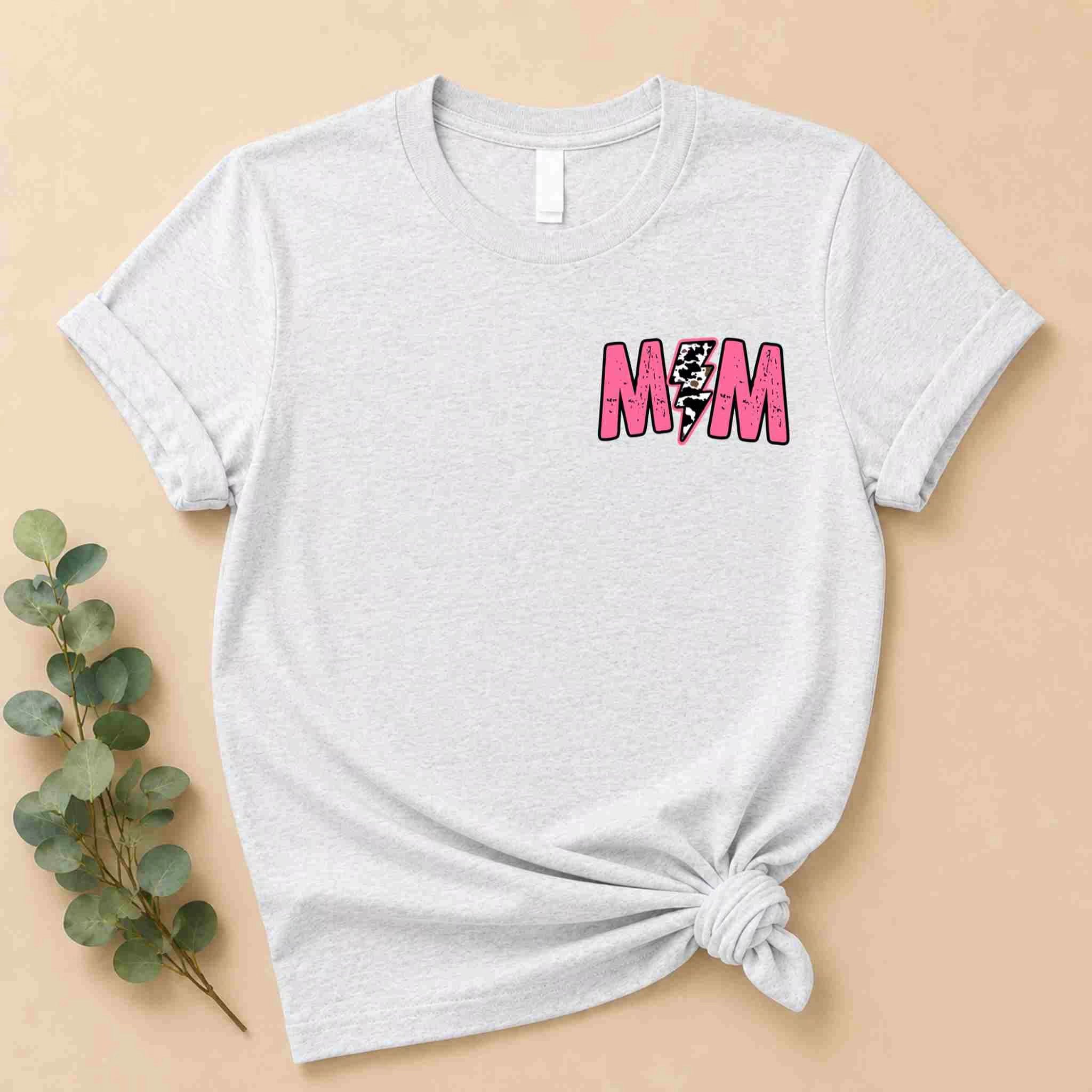 The Hot Mom Summer Tee – Trendy Mama Graphic Shirt by Becca's Banging Designs LLC is a light gray tee with bold pink M♡M and a leopard heart on the chest, knotted at the bottom, shown on beige with green eucalyptus leaves.