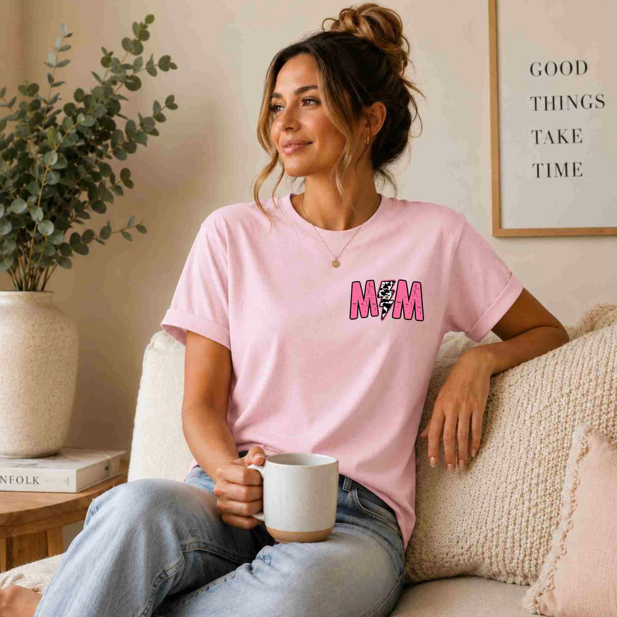 A woman with light brown hair in a bun, wearing the Hot Mom Summer Tee – Trendy Mama Graphic Shirt by Becca's Banging Designs LLC and jeans, sits on a couch holding a mug. A plant and “GOOD THINGS TAKE TIME” print are in the background.