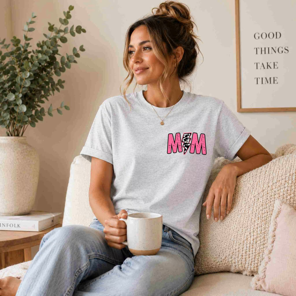A woman with a loose bun smiles while holding a mug on a beige sofa. She wears Becca's Banging Designs LLC’s Hot Mom Summer Tee—a trendy gray graphic shirt with MOM in pink—and light blue jeans. A plant and framed quote are behind her.