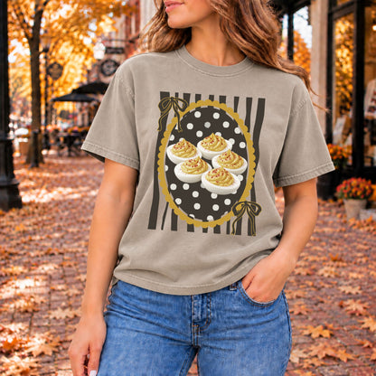 Warm gray graphic tee with five deviled eggs in a gold scalloped frame, black stripes, and gold bows.