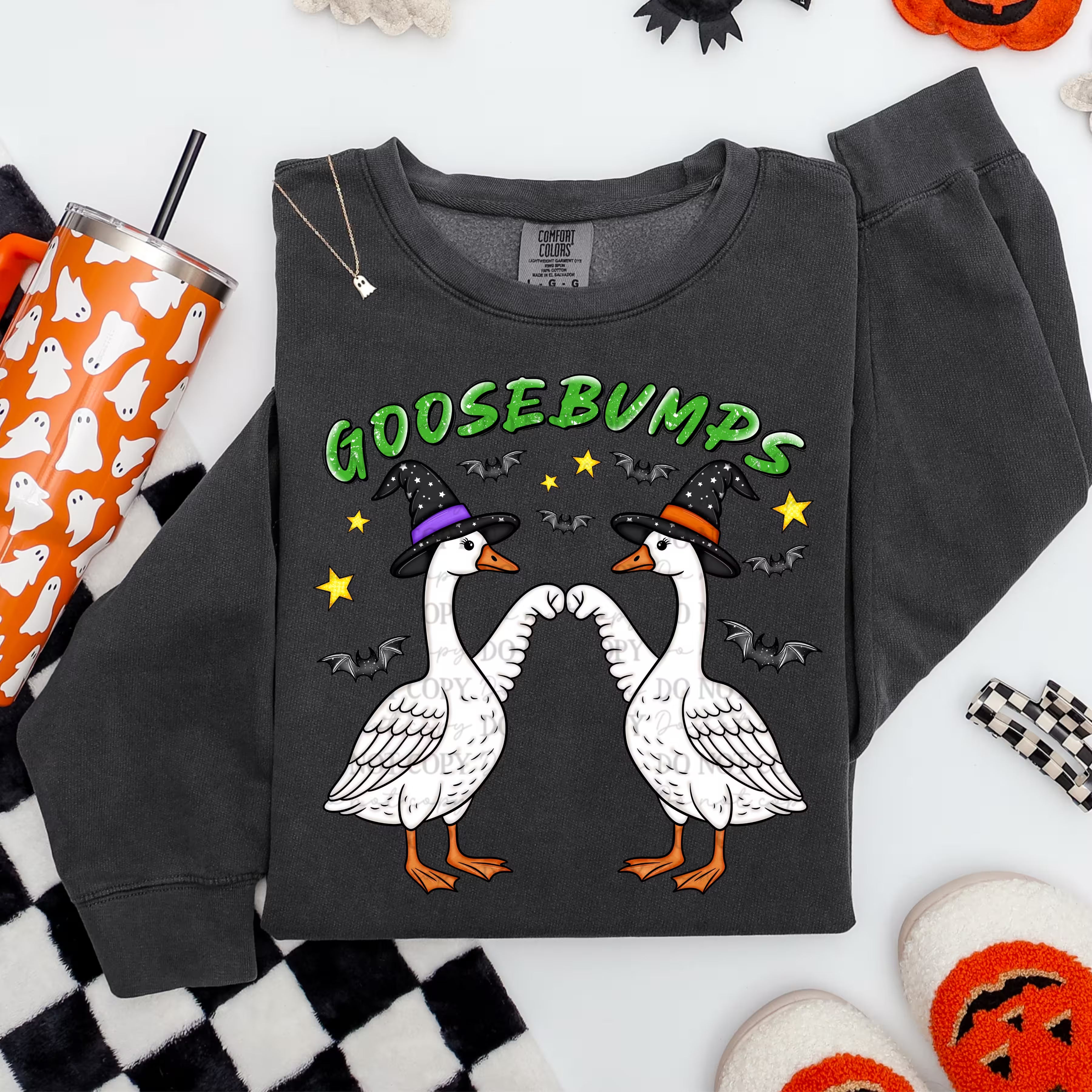 A dark charcoal gray crewneck sweatshirt featuring a Halloween graphic of two white geese wearing witch hats and facing each other, with the word 'GOOSEBUMPS' in green above them. The sweatshirt is styled in a flat lay with seasonal props including a patterned tumbler, a black and white checkered blanket, and orange pumpkin decorations.