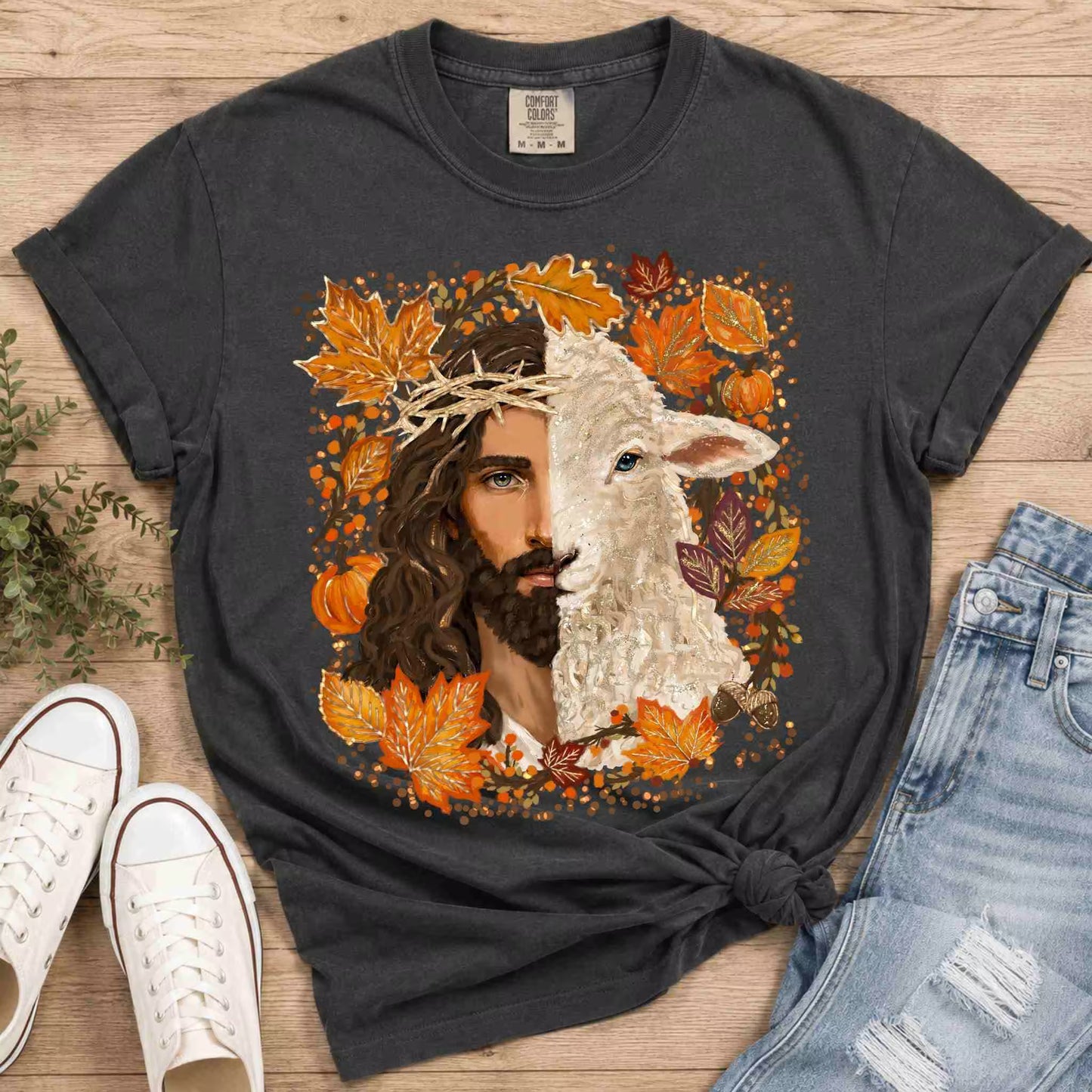 Black t-shirt with a graphic design of a man and lamb surrounded by autumn leaves and pumpkins, placed on a wooden surface with shoes and jeans.