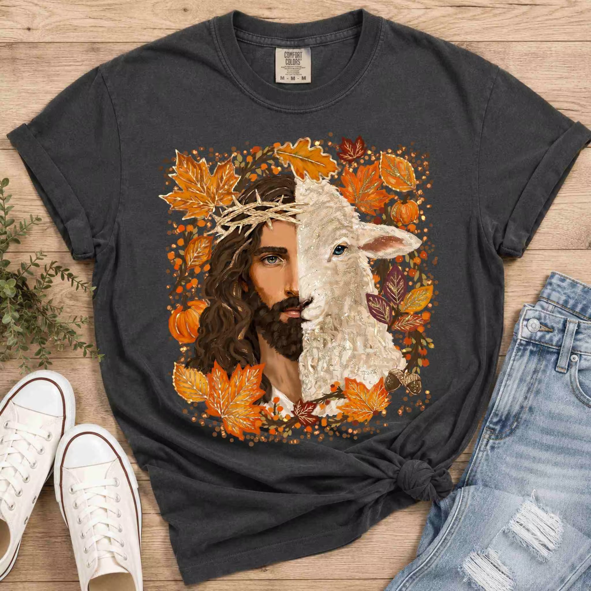 Black t-shirt with a graphic design of a man and lamb surrounded by autumn leaves and pumpkins, placed on a wooden surface with shoes and jeans.