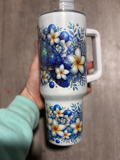 A hand holds Becca's Banging Designs LLC’s “Just a Girl Who Loves Turtles” 40oz glitter tumbler, featuring sea turtle and floral beach designs with a white handle, set against a wooden background.