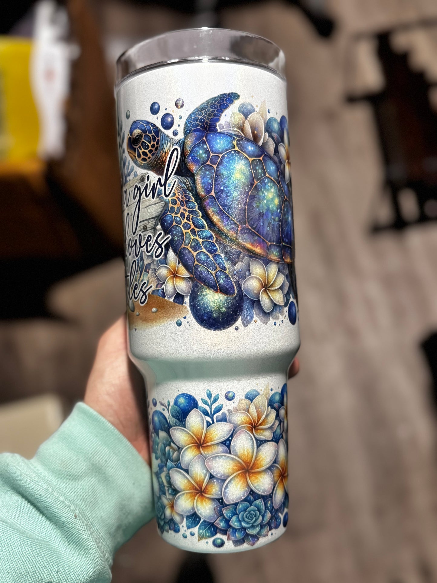A hand holds the “Just a Girl Who Loves Turtles” 40oz Glitter Tumbler by Becca’s Banging Designs LLC, featuring a blue sea turtle, plumeria flowers, ocean-themed art, and cursive text on an aesthetic handled cup.