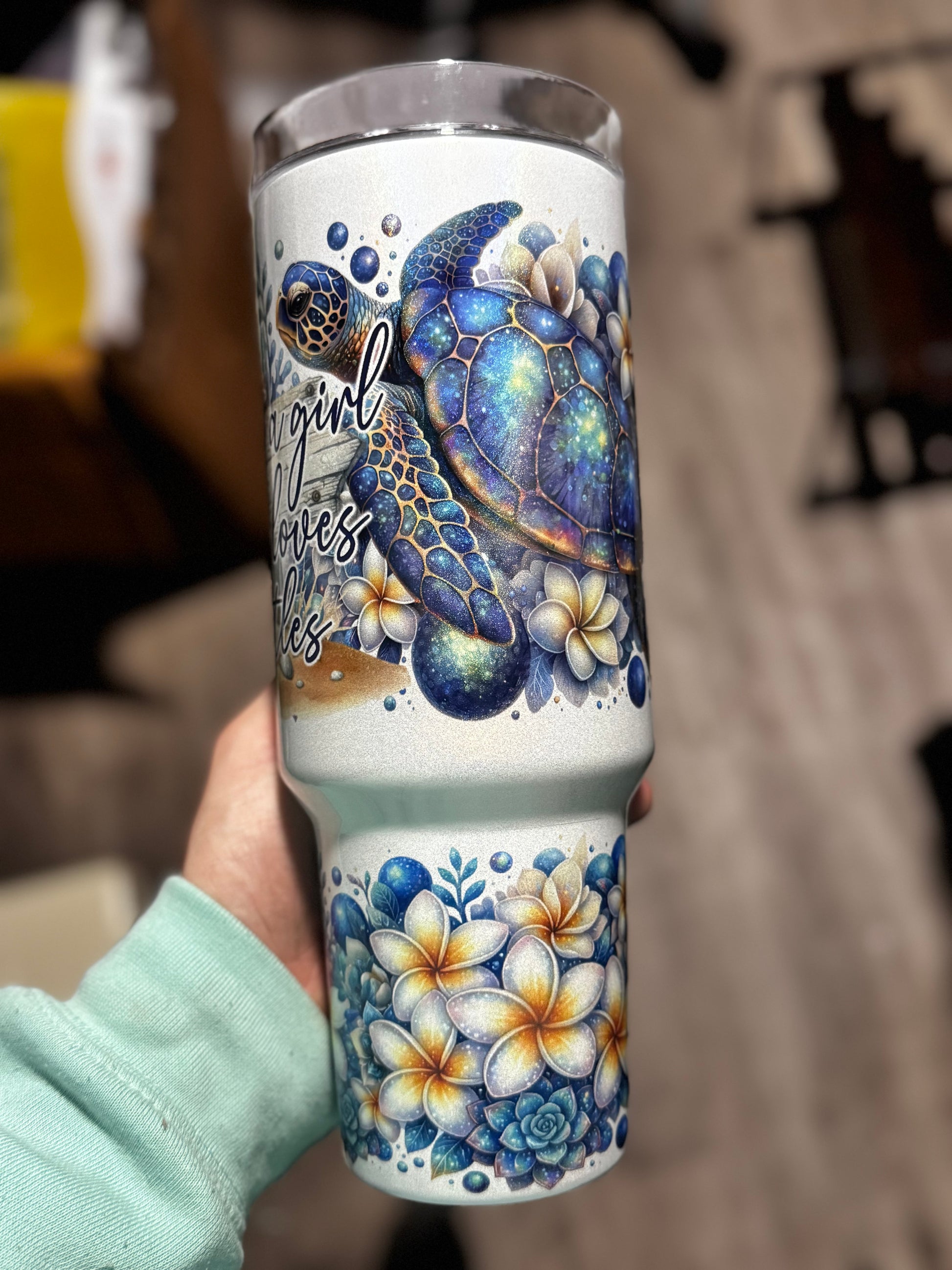 A hand holds the “Just a Girl Who Loves Turtles” 40oz Glitter Tumbler by Becca’s Banging Designs LLC, featuring a blue sea turtle, plumeria flowers, ocean-themed art, and cursive text on an aesthetic handled cup.