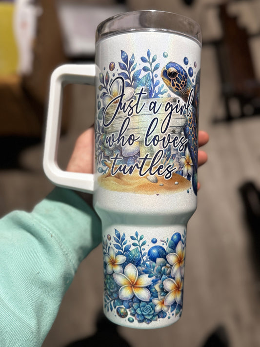 A hand holds the “Just a Girl Who Loves Turtles” Glitter Tumbler by Becca's Banging Designs LLC, a 40oz cup with handle featuring sea turtles and blue and yellow floral designs. The background is blurred.