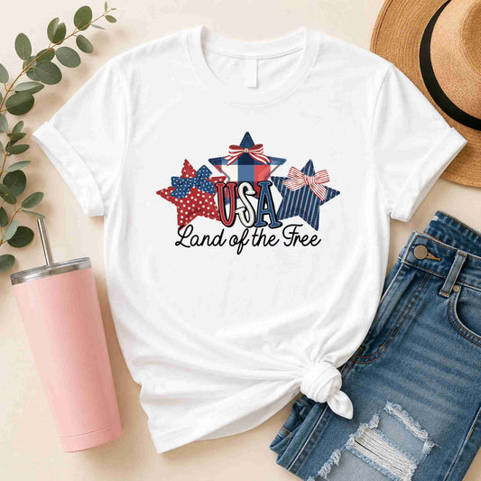 The Land of the Free USA T-Shirt by Becca's Banging Designs LLC features patriotic bow star graphics, styled with ripped denim shorts, a hat, a pink tumbler, and greenery for a festive red, white, and blue look.