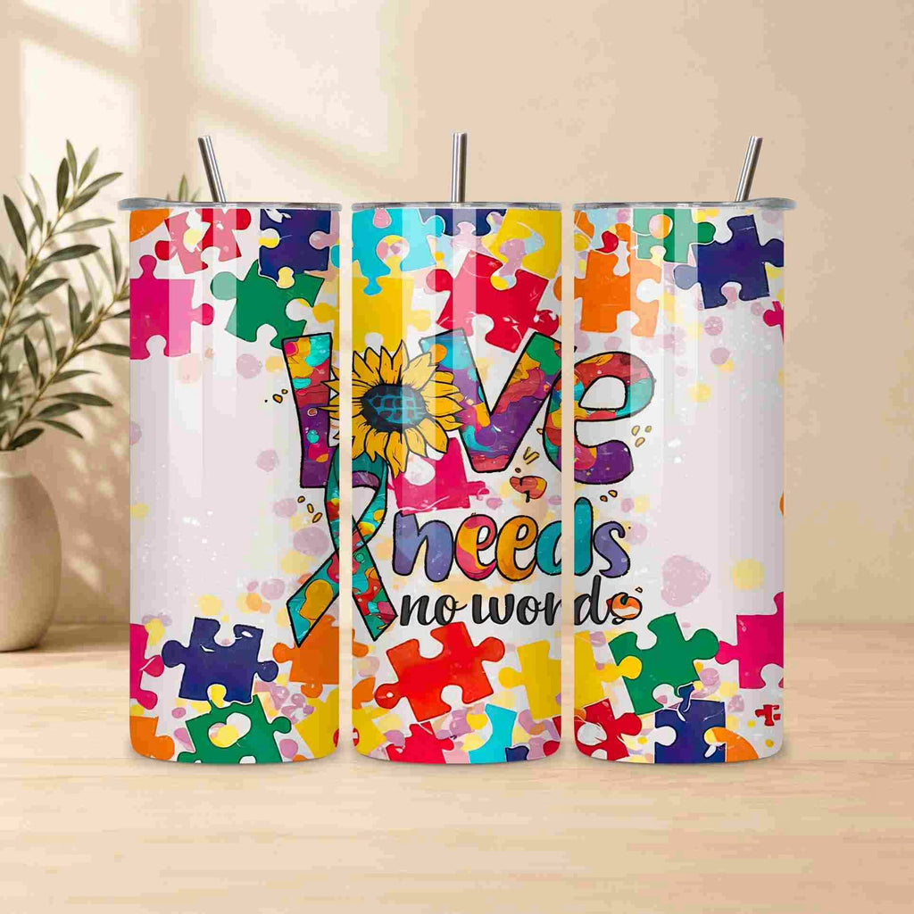 The Autism Awareness Love Needs No Words Tumbler by Becca's Banging Designs LLC features a puzzle piece sunflower and ribbon design—an ideal autism support gift—displayed on a light wood surface with a plant in the background.