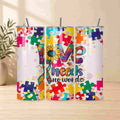 The Autism Awareness Love Needs No Words Tumbler by Becca's Banging Designs LLC features a puzzle piece sunflower and ribbon design—an ideal autism support gift—displayed on a light wood surface with a plant in the background.