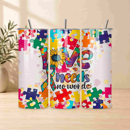 The Autism Awareness Love Needs No Words Tumbler by Becca's Banging Designs LLC features a puzzle piece sunflower and ribbon design—an ideal autism support gift—displayed on a light wood surface with a plant in the background.