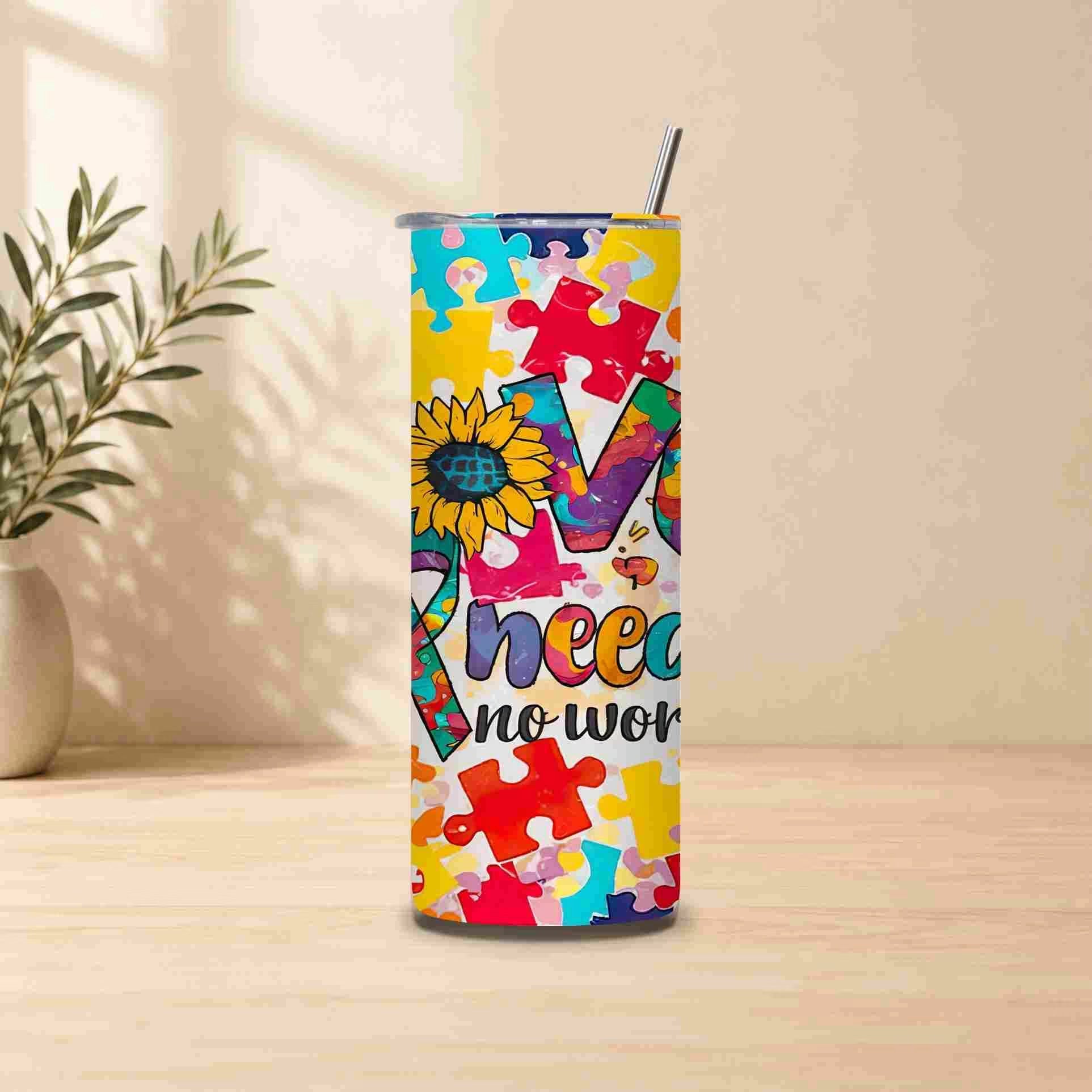 The Autism Awareness Love Needs No Words Tumbler by Becca's Banging Designs LLC features colorful puzzle pieces, a sunflower, and an awareness ribbon. This 20oz cup is an inspiring autism support gift. Displayed on a wooden table with a plant nearby.