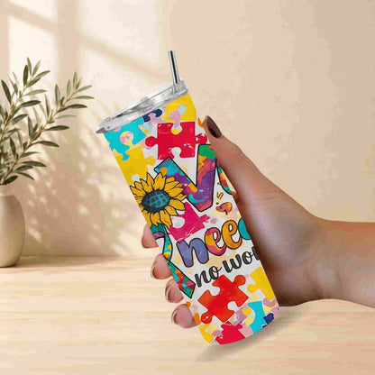 A hand holds Becca's Banging Designs LLC Autism Awareness Love Needs No Words Tumbler, a 20oz cup featuring colorful puzzle pieces and a sunflower, set against a light background with a plant in the distance.