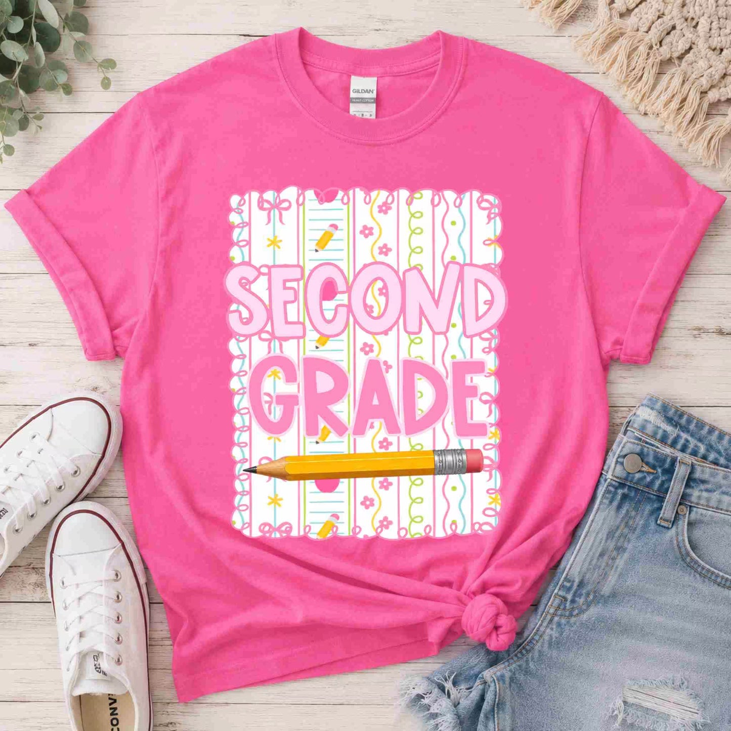 Pink t-shirt with 'SECOND GRADE' text and pencil graphic on a wooden surface with shoes and jeans.