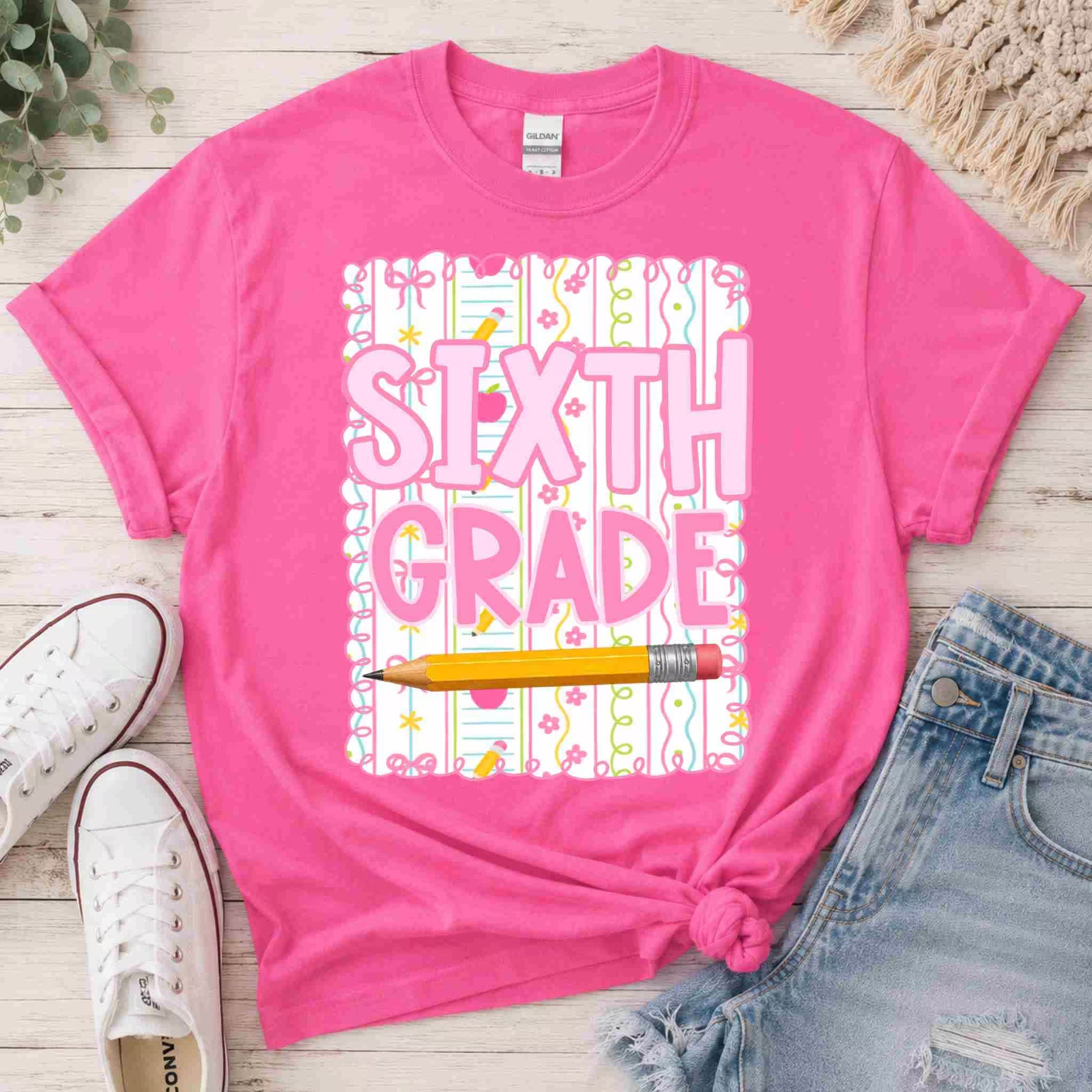Pink t-shirt with 'Third Grade' text and pencil graphic on a wooden surface with shoes and jeans.