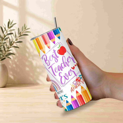 A hand holds the Best Teacher Ever | 20oz Tumbler, decorated with pencils, apples, and words like #teacher, INSPIRE, and TEACH. A straw is in the lid. A small plant sits blurred in the background—a great teacher gift.