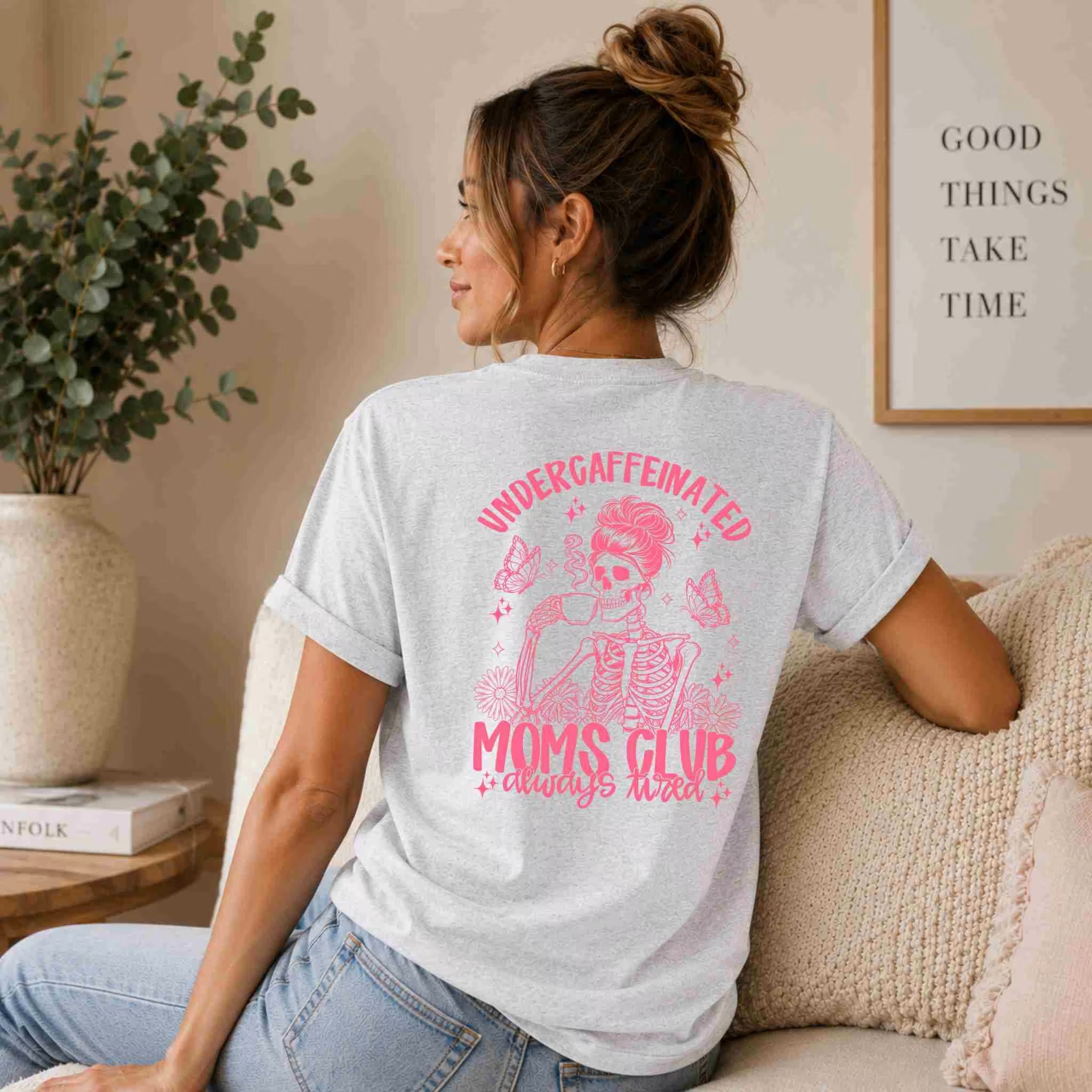 A woman with a messy bun sits on a couch, rocking the Undercaffeinated Moms Club Shirt – Funny Tired Mom Tee from Becca's Banging Designs LLC, which features a pink skeleton graphic. A plant and framed sign accent the cozy backdrop.