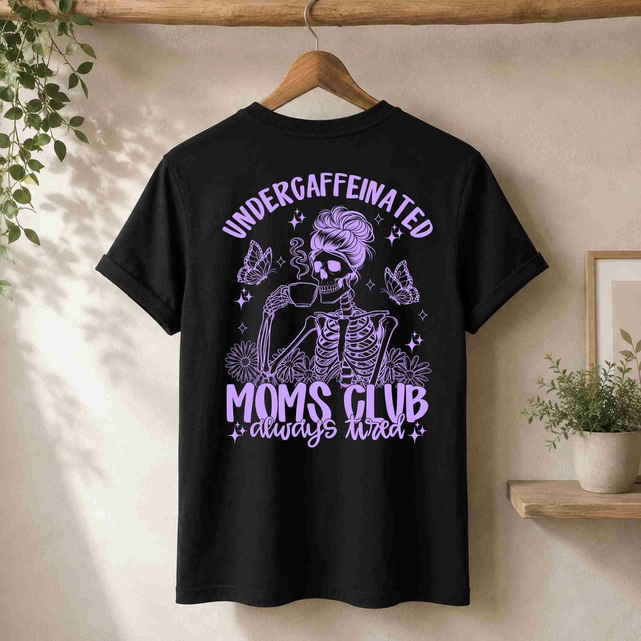 A black Undercaffeinated Moms Club Shirt by Becca's Banging Designs LLC hangs on a wooden hanger, with “ALWAYS TIRED” and a purple skeleton mom graphic—ideal for cute t shirt outfits and fans of funny tired mom tees.