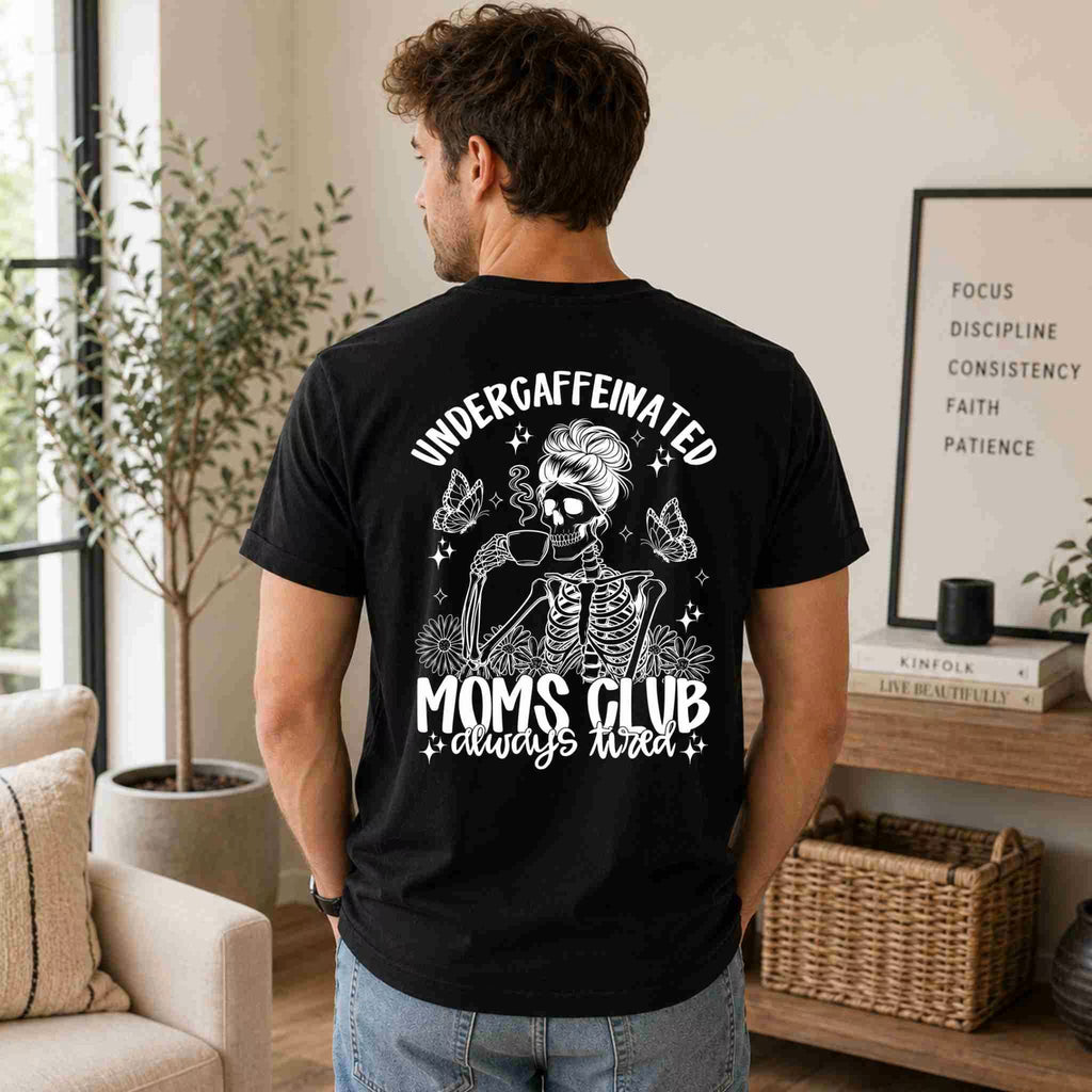 A man indoors wears Becca's Banging Designs LLC’s Undercaffeinated Moms Club Shirt—a black women’s tee with a skeleton mom in sunglasses and a bun sipping coffee, reading: Undercaffeinated Moms Club, always tired.