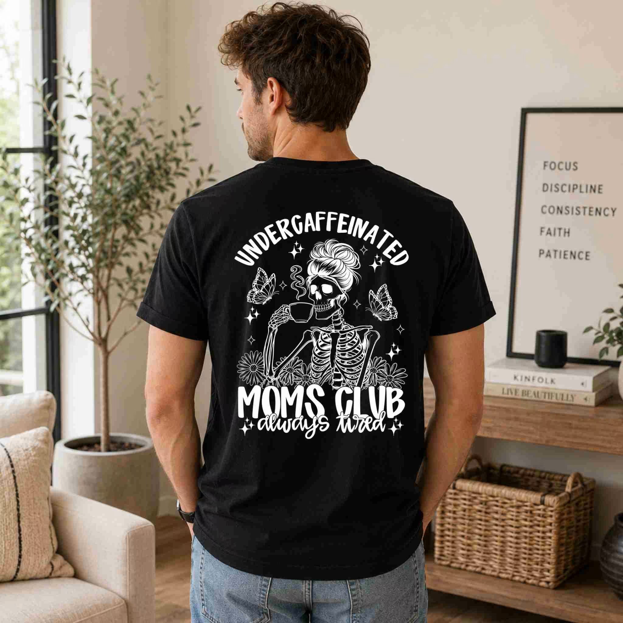 A man indoors wears Becca's Banging Designs LLC’s Undercaffeinated Moms Club Shirt—a black women’s tee with a skeleton mom in sunglasses and a bun sipping coffee, reading: Undercaffeinated Moms Club, always tired.
