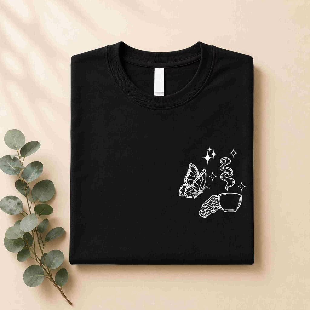 The Undercaffeinated Moms Club Shirt by Becca's Banging Designs LLC is a folded black tee with white line art of a butterfly, hand, sparkles, and steaming cup—perfect for cute outfits for tired moms. Placed on beige with eucalyptus leaves.