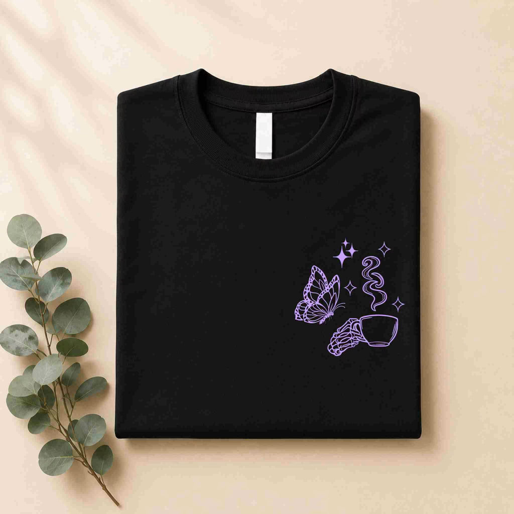 A folded black Undercaffeinated Moms Club Shirt by Becca's Banging Designs LLC features a purple line art butterfly, a hand with a steaming cup, and sparkles—perfect for cute t-shirt outfits. Displayed on beige next to eucalyptus.