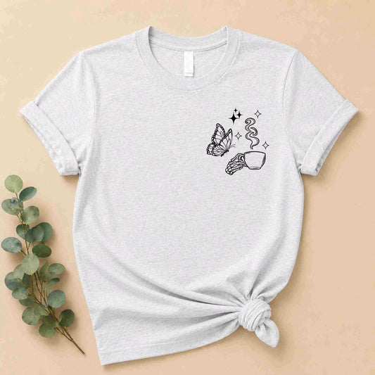 The Undercaffeinated Moms Club Shirt by Becca’s Banging Designs LLC is a light gray women’s tee featuring black line art of a butterfly, a hand with a steaming cup, and stars—perfect for cute knotted t-shirt looks beside green leaves.