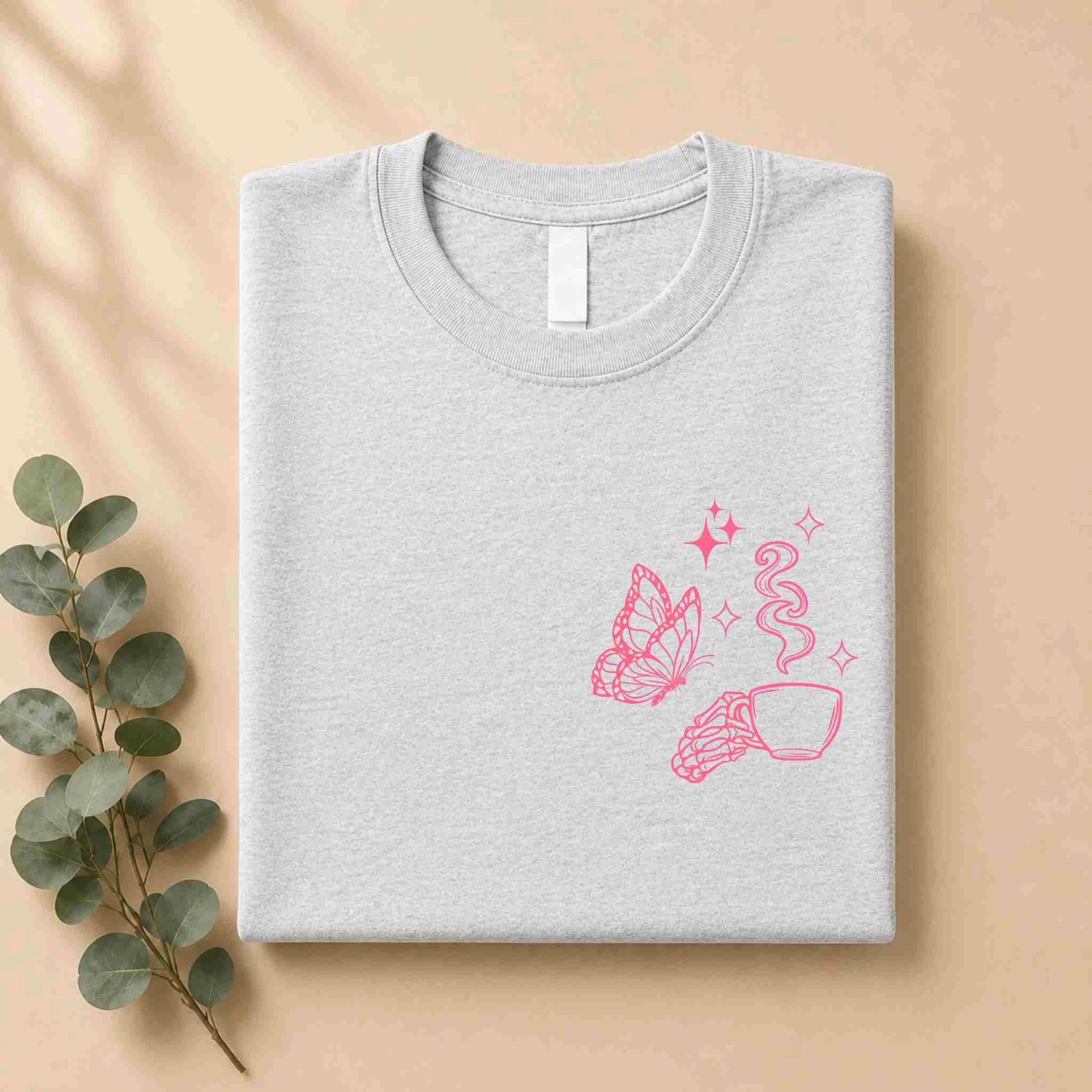 The Undercaffeinated Moms Club Shirt – Funny Tired Mom Tee by Becca's Banging Designs LLC features a pink butterfly, coffee cup, and croissant on light gray. Folded on a beige surface with eucalyptus, it’s perfect for cute t shirt outfits.