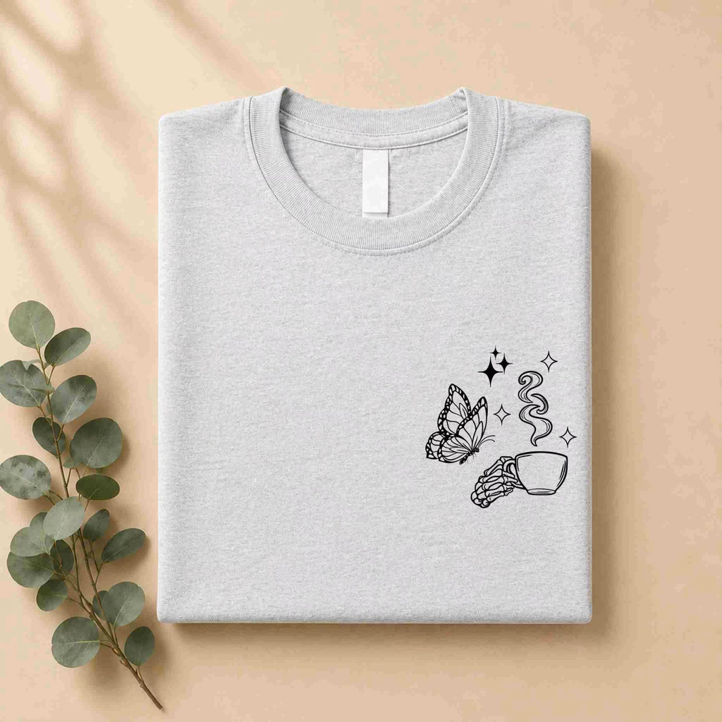 A folded light gray Undercaffeinated Moms Club Shirt by Becca's Banging Designs LLC features black line art of a butterfly, a hand with a steaming cup, and small stars—perfect for cute t shirt outfits. It is styled on beige with eucalyptus beside it.