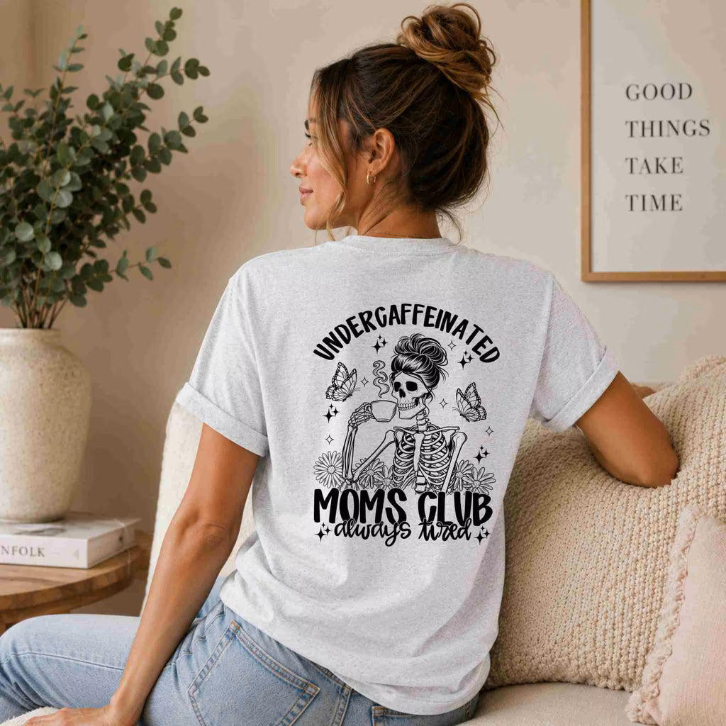A woman with her hair in a bun relaxes on a couch, wearing the Undercaffeinated Moms Club Shirt – Funny Tired Mom Tee by Becca's Banging Designs LLC. A "GOOD THINGS TAKE TIME" sign behind her completes this charming mom outfit moment.