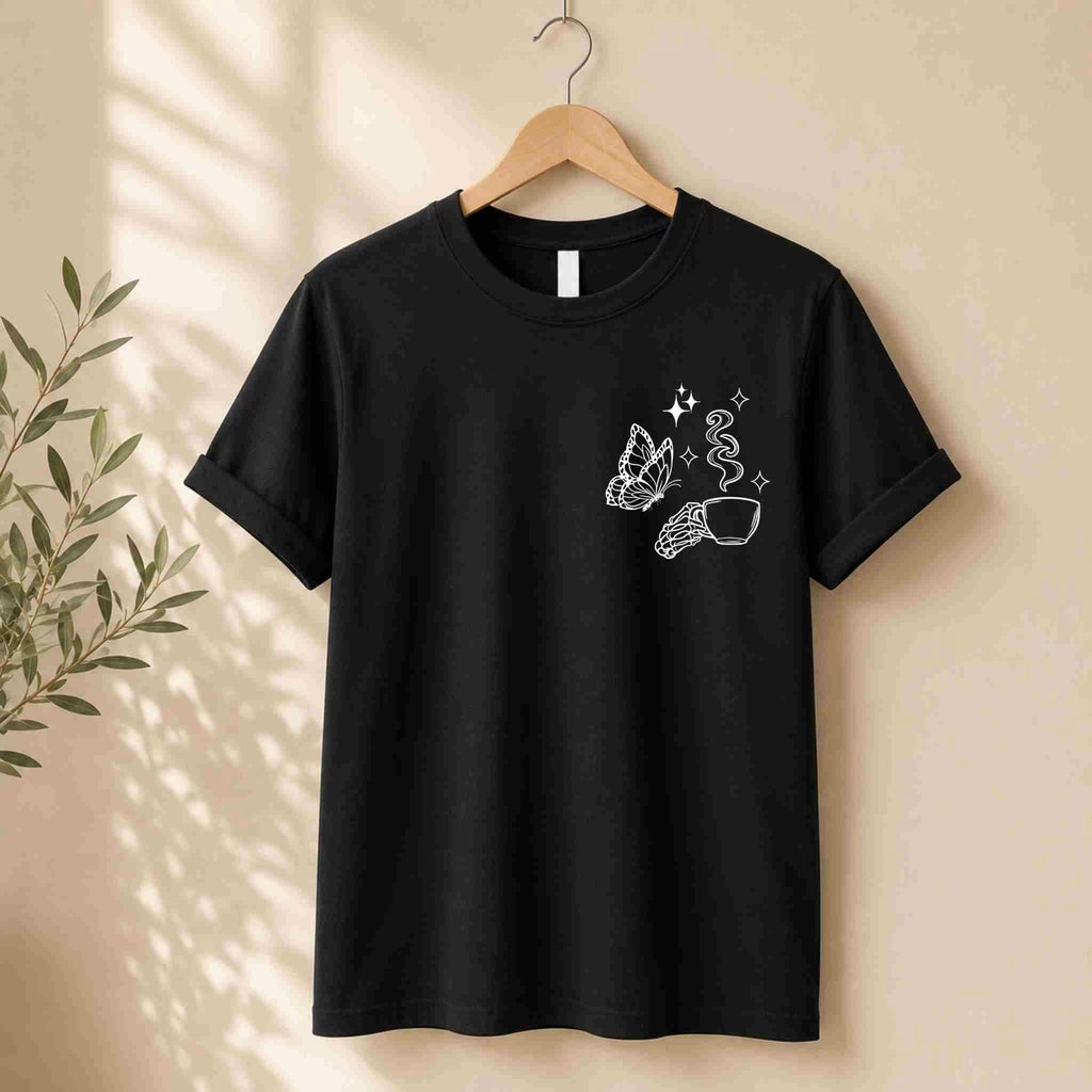 The Undercaffeinated Moms Club Shirt – Funny Tired Mom Tee by Becca's Banging Designs LLC hangs on a wooden hanger, showing white butterfly, hand, sparkles, and steaming cup art—ideal for cute t-shirt outfits. Beige wall and leafy plant nearby.