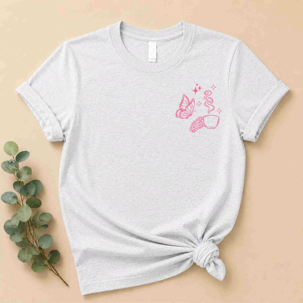 The Undercaffeinated Moms Club Shirt by Becca's Banging Designs LLC is a light gray women's graphic tee with a pink butterfly, sparkles, and a hand holding a sprouting plant. Styled with a knotted hem and displayed on beige with green leaves.