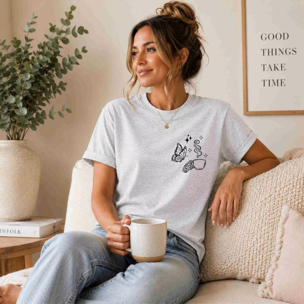A woman with her hair in a bun sits on a couch holding a mug. She wears the "Undercaffeinated Moms Club Shirt" by Becca's Banging Designs LLC with blue jeans. A potted plant and “GOOD THINGS TAKE TIME” wall sign are behind her.