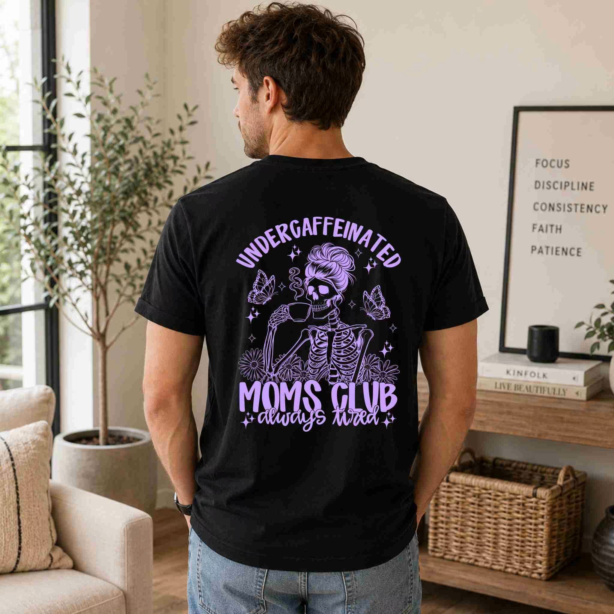 A man stands indoors in a cozy living room wearing Becca's Banging Designs LLC's Undercaffeinated Moms Club Shirt – Funny Tired Mom Tee, featuring a skeleton mom graphic and the "always tired" slogan in purple and white.
