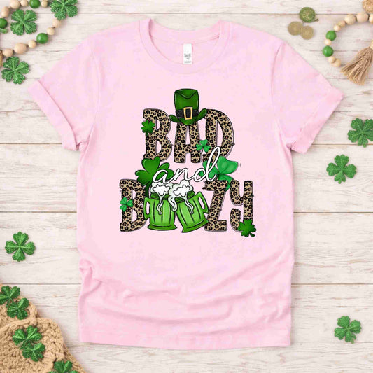 St. Patricks Day shirt featuring 'Bad and Boozy' graphic with leopard print, shamrocks, and beer mugs on light pink tee.