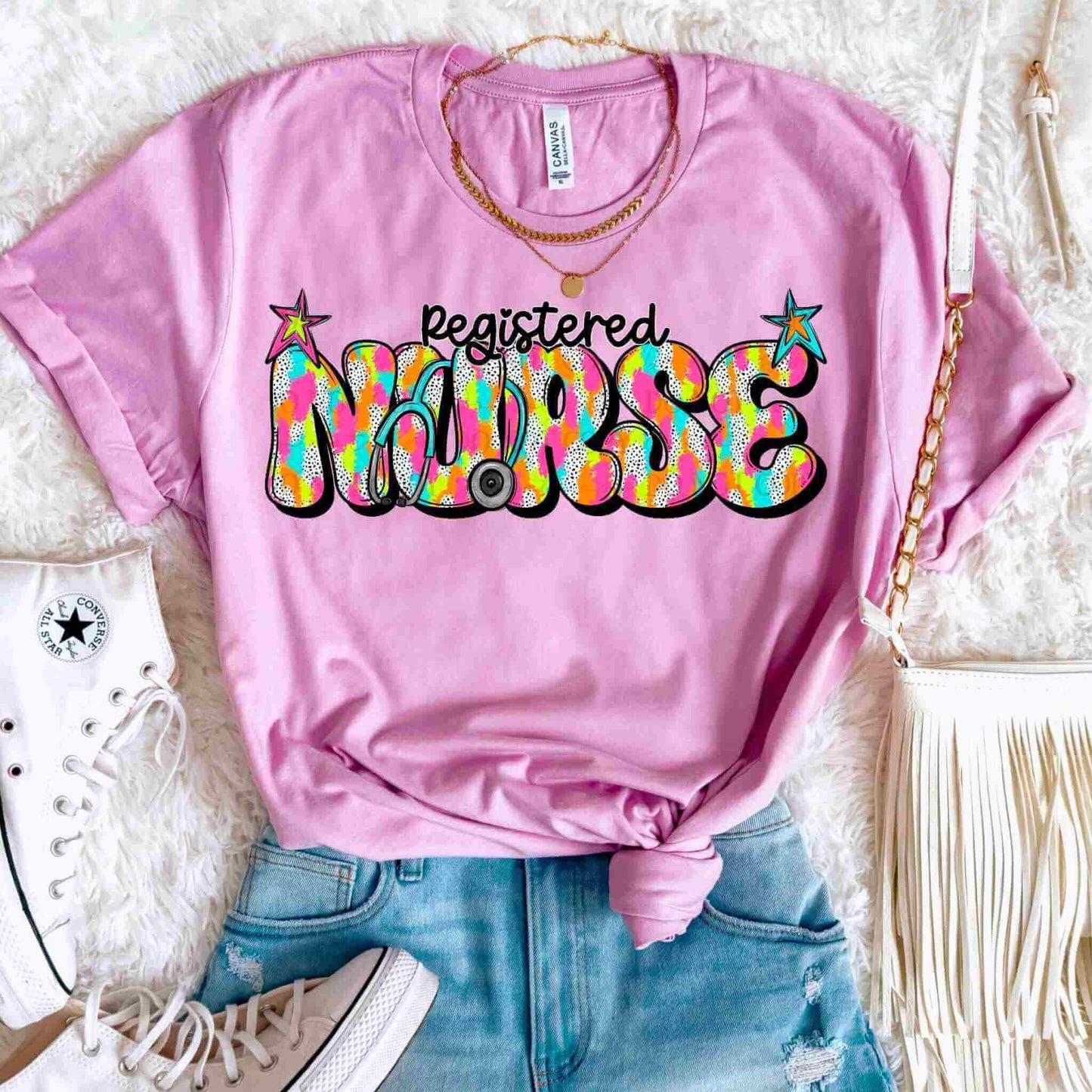 A Colorful Nurse Shirt – Registered Nurse by Becca's Banging Designs LLC is styled with ripped denim shorts, white Converse shoes, layered gold necklaces, and a white fringed bag on a fuzzy white surface.