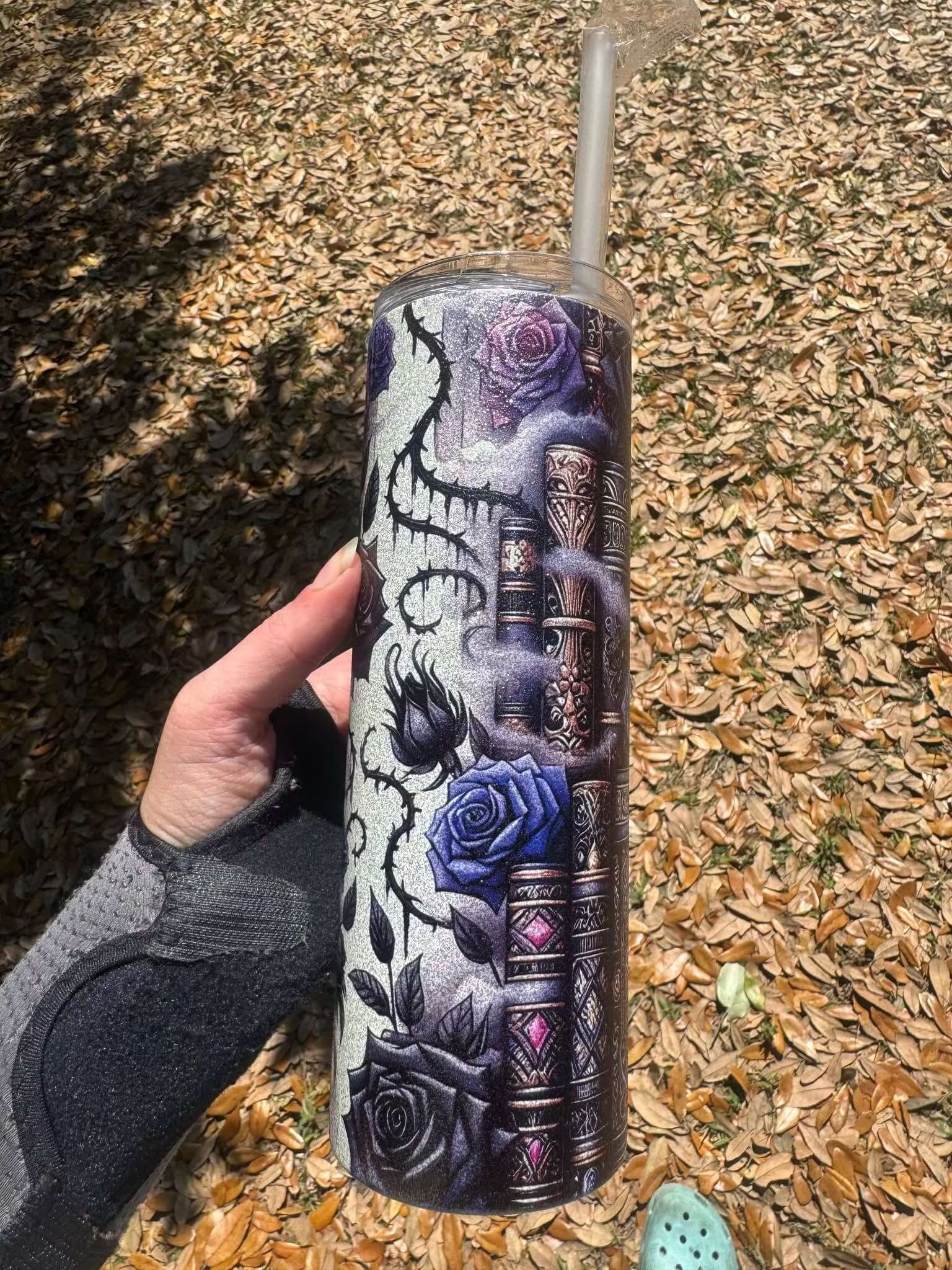 A hand in a wrist brace holds the Dark Book Lover 20oz Skinny Tumbler by Becca's Banging Designs LLC, featuring purple and black roses, a spider, and gothic patterns. Dry brown leaves and a green shoe are visible in the background.