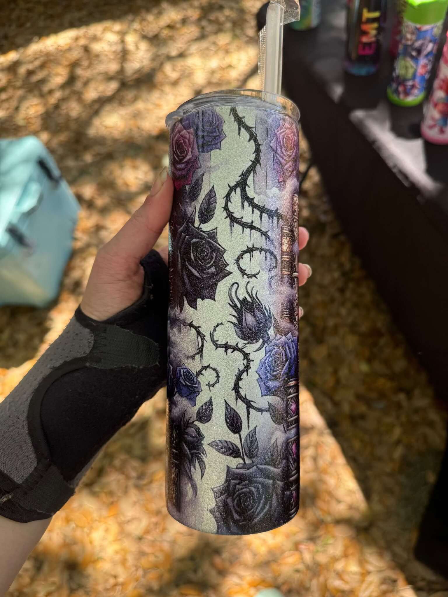 A hand in a black wrist brace holds the Dark Book Lover 20oz Skinny Tumbler by Becca's Banging Designs LLC, decorated with dark purple and black roses, thorny vines, and a straw. Autumn leaves are scattered on the ground below.