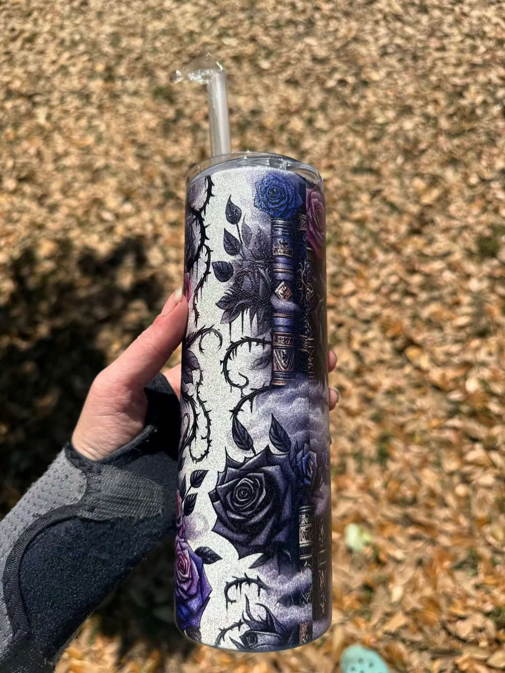 A hand holds the Dark Book Lover 20oz Skinny Tumbler by Becca's Banging Designs LLC, featuring dark purple and blue roses, thorny vines, and books, outdoors above a layer of dry leaves. Comes with lid and straw.