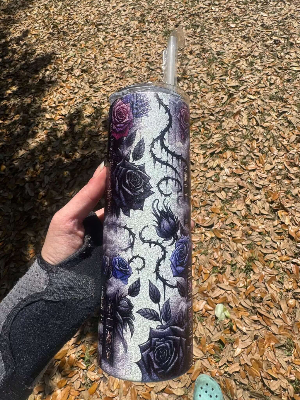 A hand in a wrist brace holds the Dark Book Lover 20oz Skinny Tumbler by Becca's Banging Designs LLC—glittery with black and purple roses. Fallen brown leaves cover the ground, and a green shoe peeks in at the bottom right.