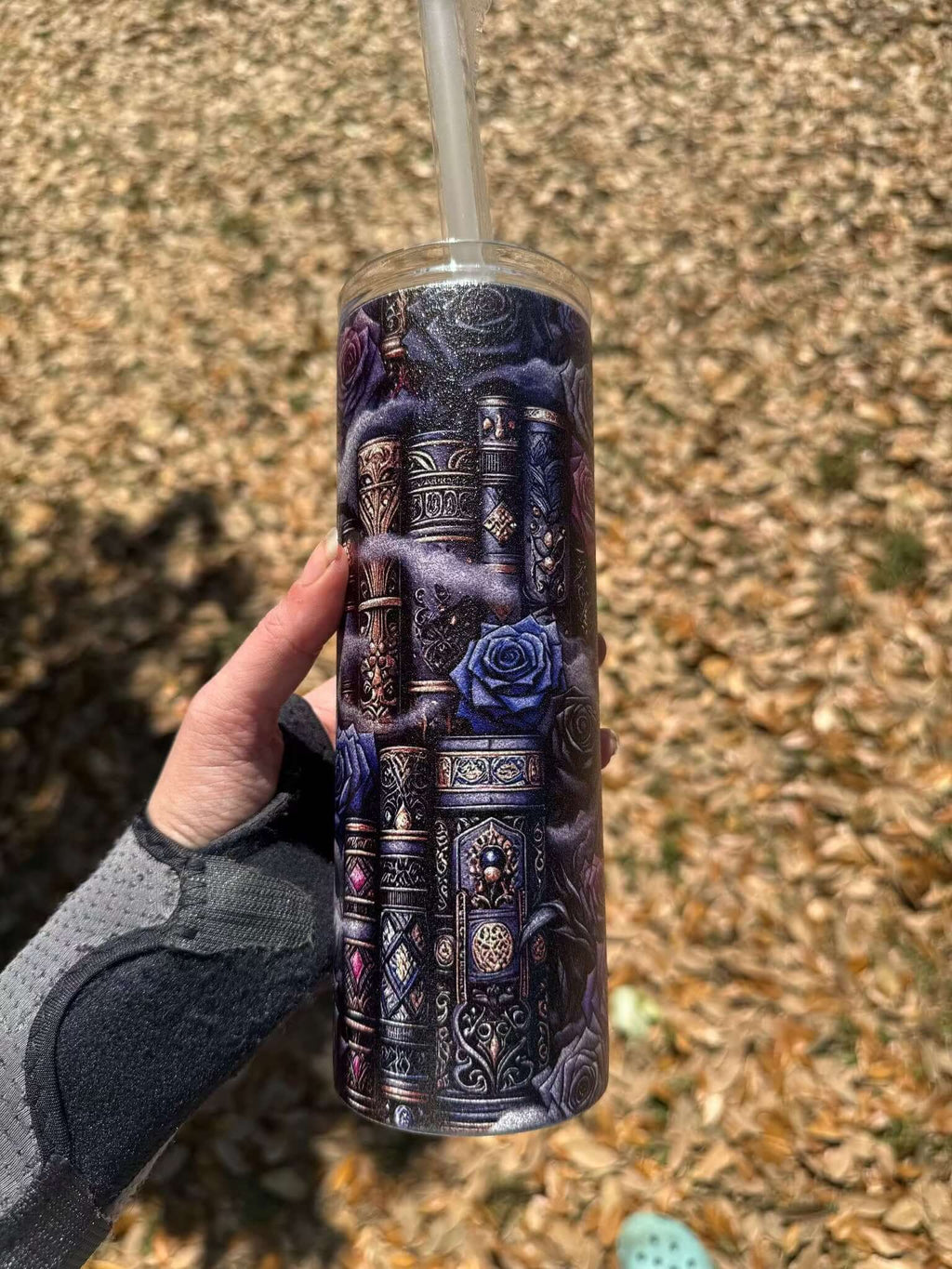 A hand holds the Dark Book Lover 20oz Skinny Tumbler by Becca's Banging Designs LLC, featuring ornate book spines and blue roses, with dry leaves scattered on the ground in the background.