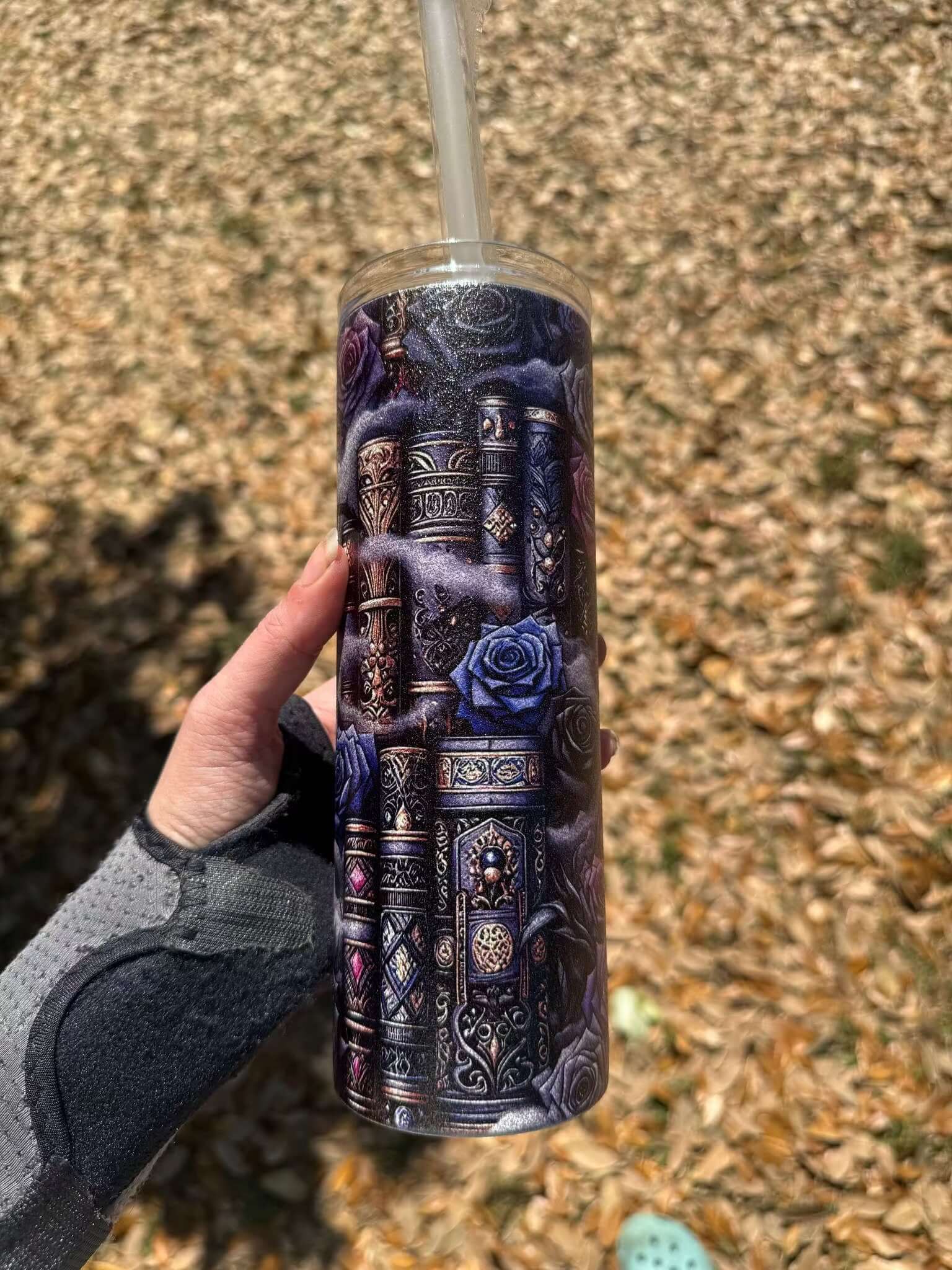 A hand holds the Dark Book Lover 20oz Skinny Tumbler by Becca's Banging Designs LLC, featuring ornate book spines and blue roses, with dry leaves scattered on the ground in the background.
