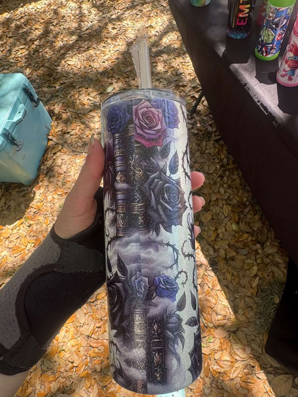 A hand holds the Dark Book Lover 20oz Skinny Tumbler by Becca's Banging Designs LLC, featuring dark and purple roses, gothic books, and a cloudy sky. Colorful tumblers and scattered leaves are visible in the background.