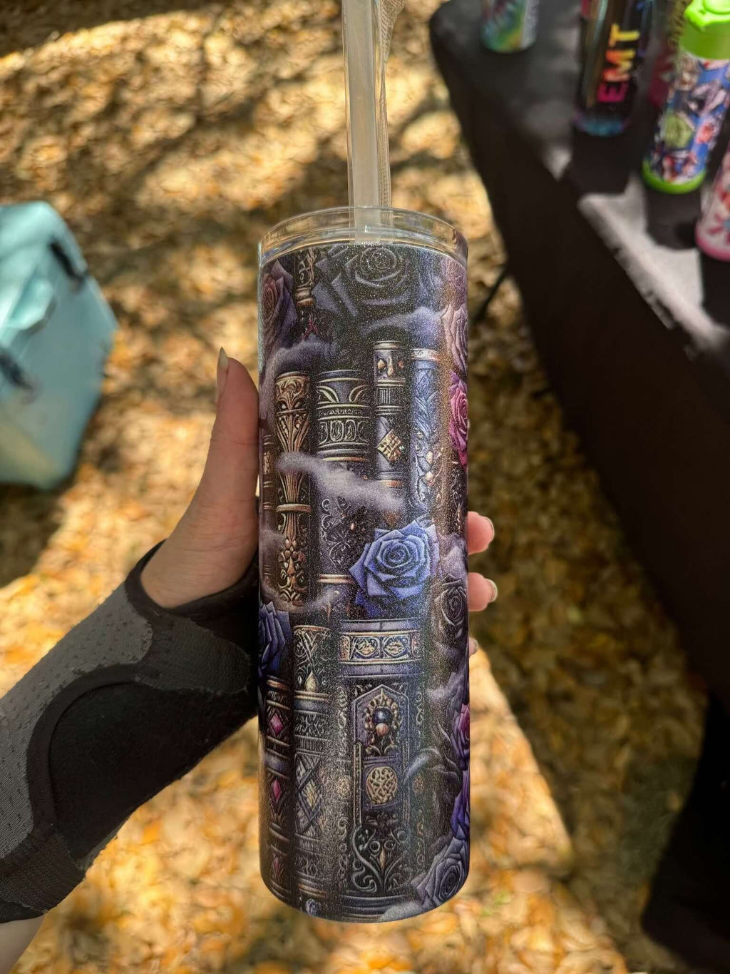 A hand holds the Dark Book Lover 20oz Skinny Tumbler by Becca's Banging Designs LLC, featuring ornate silver patterns and purple roses. In the shaded outdoor background, fallen leaves and other colorful tumblers sit on the table.